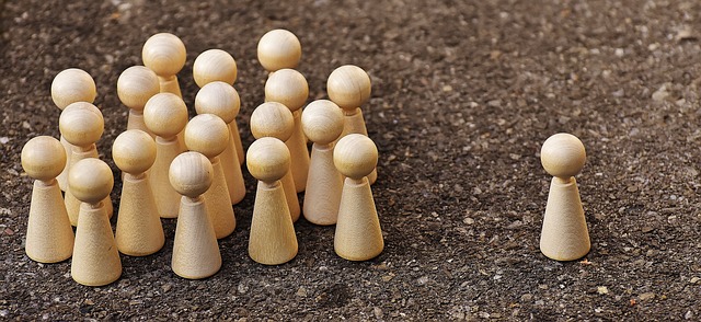Image Description: a picture displaying a group of pale-coloured wooden pawns all amassed into a group, with the exception of one pawn outside the circle to the left. The larger group of pawns are facing the single pawn as a collective.