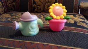 Image Description: two small bath toys, left to right, a purple and robin's egg blue water-can and a a yellow and pink flower with a bright-pink pot. you can use them to blow at people or yourself.