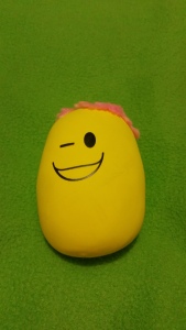 Image Description: a small rubbery yellow stress-ball with a winky face and pink woollen hair