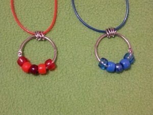 Image Description: two bead-ring necklaces on a red Rainbow String and a dark-blue Rainbow String with four chrome loops in between the Rainbow String and the key-ring/split ring. From left to right, a red themed bead-ring necklace (transparent red, acrylic red, pearlized cranberry-red, acrylic red, transparent red) and a blue themed bead-ring necklace (transparent dark-blue, acrylic dark-blue, pearlized dark-blue, acrylic dark-blue, transparent dark-blue)