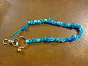 Image Description: from right to left, a chrome key-ring and lobster-claw clasp, attached to the key-ring is small gold-coloured bell. Also attached to the key ring is a long beaded lanyard made of robin's egg blue coloured elastic with with five different types of beads (acrylic, transparent, pearl, glittery, hourglass) displayed in variations of light blue with a blue-glittery-star-bead with silver glitter in between each group of beads, with a blue-glittery-star-bead with silver glitter as a stopper at the end.
