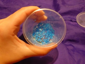 Image Description: a white hand holding a small plastic container that contains Saxa rock salt crystals that have been died blue, coated in white glitter and lavender essential oil has been added to it